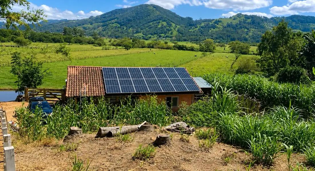 Sistema fotovoltaico no telhado de uma construção rural, com pastagens e morros ao fundo